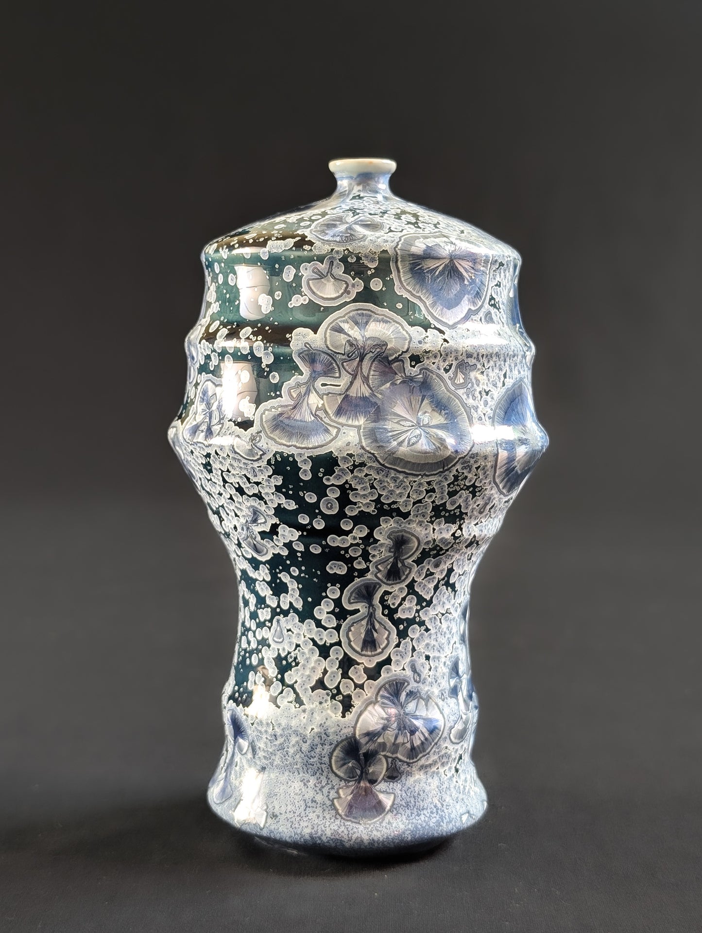 Tall, narrow-neck crystalline vase with flowing curves and a deep midnight blue glaze, covered in intricate silver and lavender crystal formations, shown against a dark background.