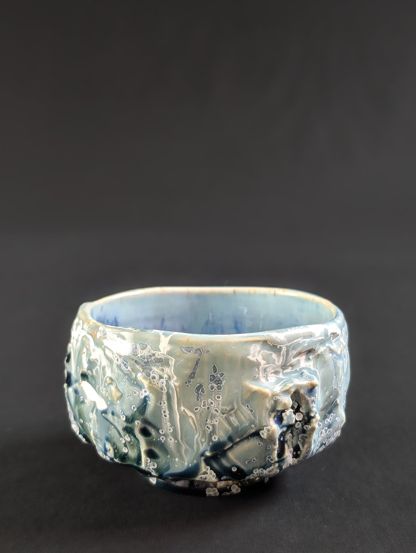 Angled profile of the chawan with glaze dripping over heavy carving, accented by tiny white-rimmed blue crystals.