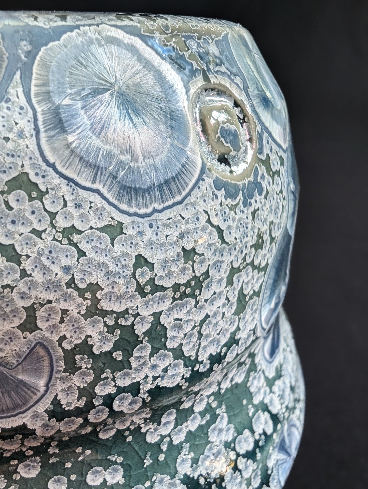 Close up shot of porcelain vase 'Earth's Jewel' showing a large sapphire crystal and many tiny silver secondary crystals against an emerald green background.