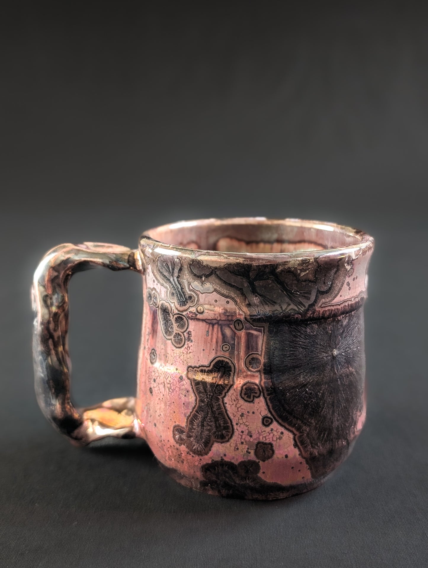 A handcrafted crystalline mug named Pink Nebula, featuring a luminous copper-pink glaze with dramatic black crystal formations rising across the surface.