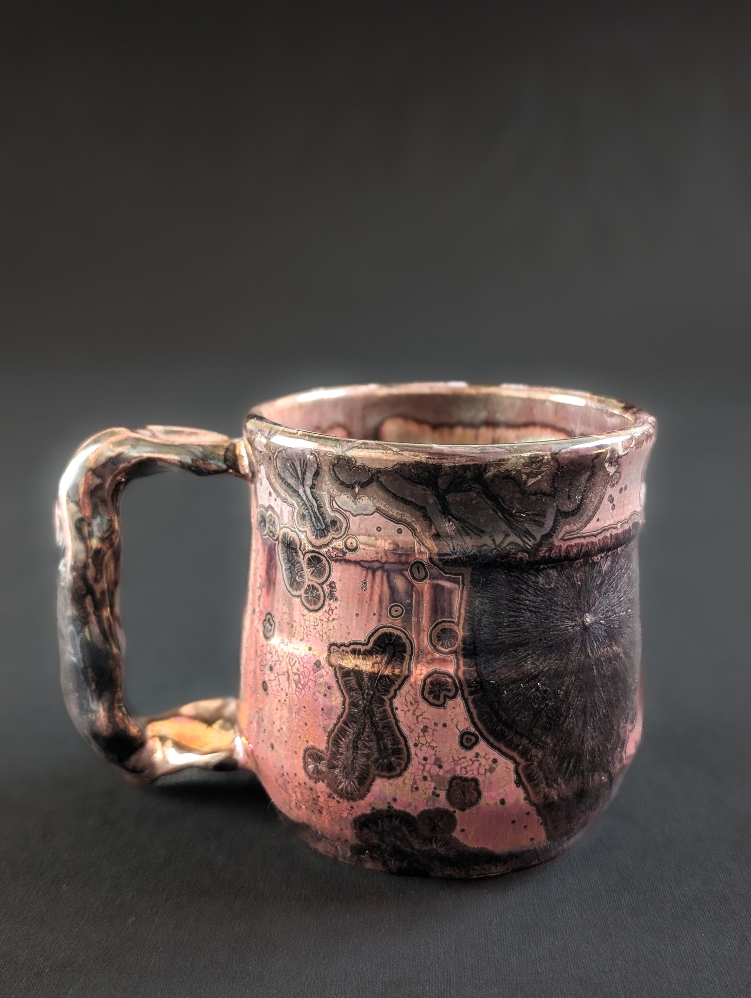 A handcrafted crystalline mug named Pink Nebula, featuring a luminous copper-pink glaze with dramatic black crystal formations rising across the surface.