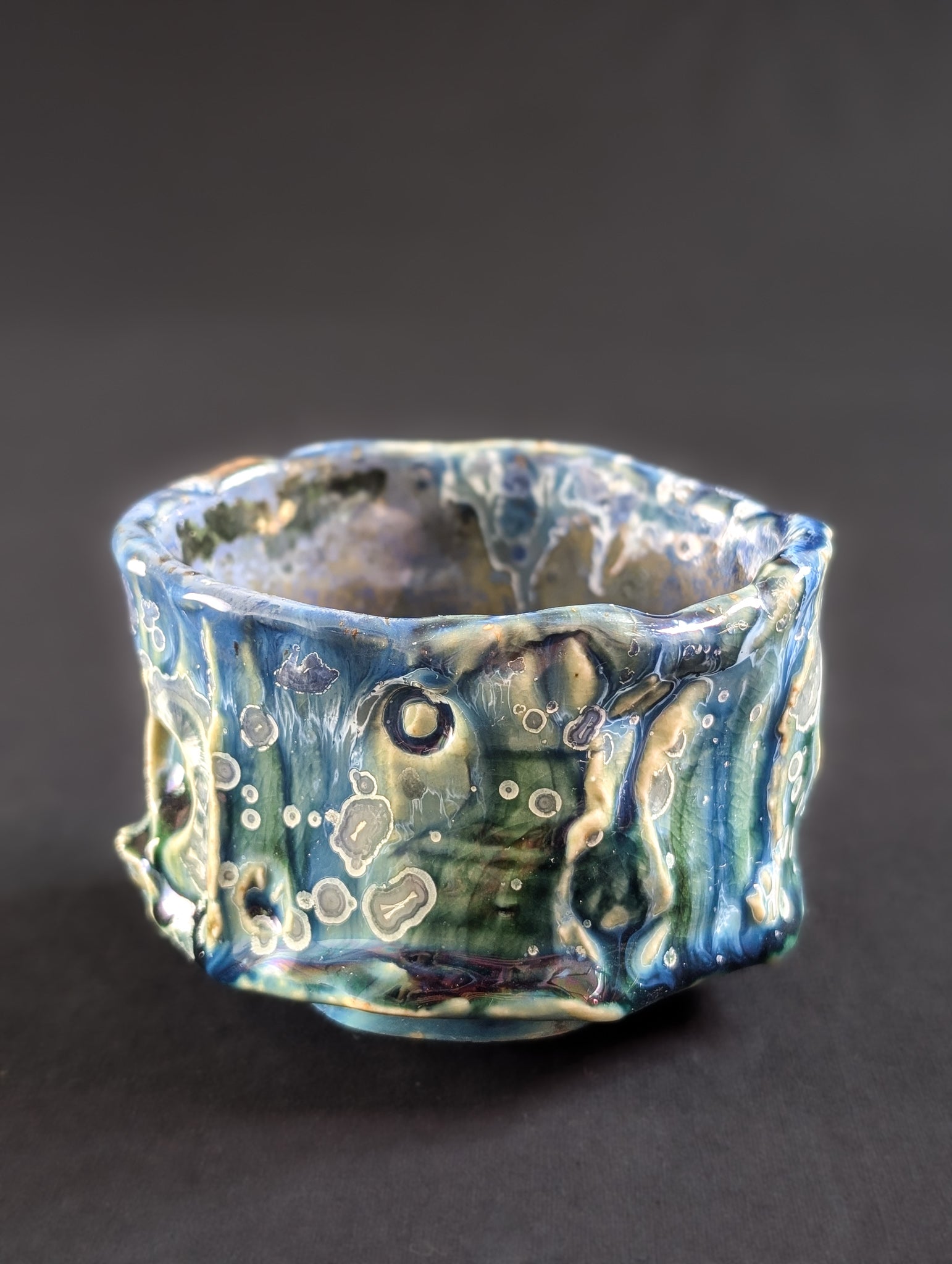 Side view of the 'Rainforest Whisper' chawan by Robbin Harris, showing deep green and blue glaze with crater-like texture and scattered yellow crystals with silver halos.