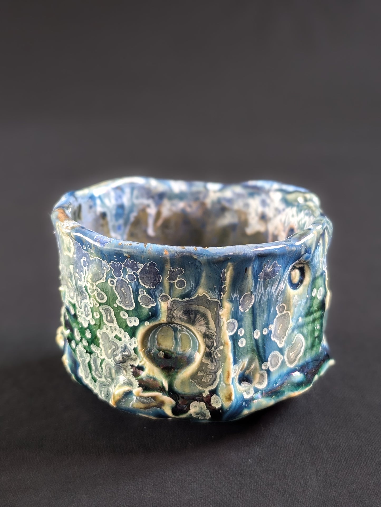 Side view of 'Rainforest Whisper' chawan with rich, layered glazes and small yellow crystals across a jungle-dark backdrop.