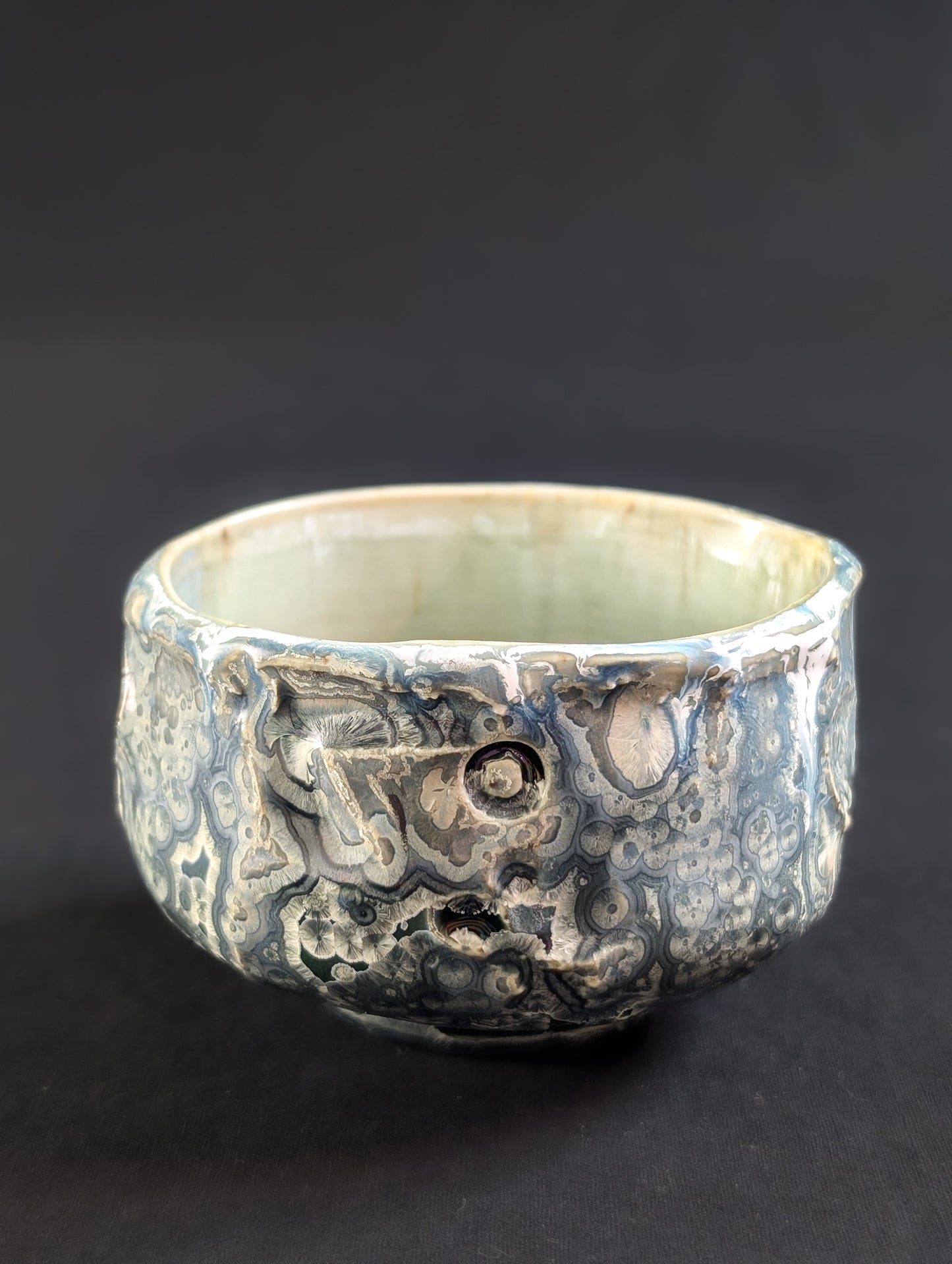 Side view of 'Tetsuya', where soft gray-blue tones and shimmering crystals emerge from a dark, elemental glaze.