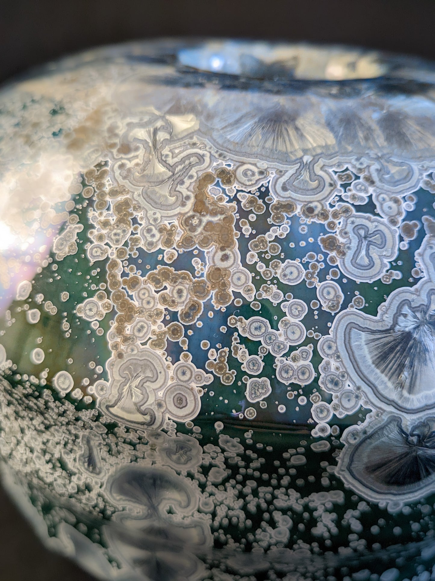 Close up of porcelain vase titled "Milky Green Cosmos," featuring a green gradient background, large deep blue crystals with small silver ones, and a sculptural cap with three parts. 