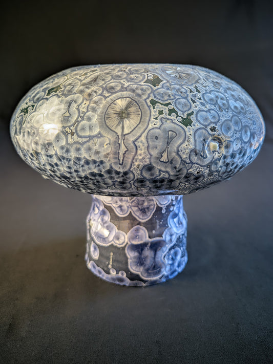 Small porcelain vase titled "Cosmic Fungi," featuring light blue and silver ringed crystals, deep blue stem, and a bulbous cap with glaze drips, set against a contrasting background.