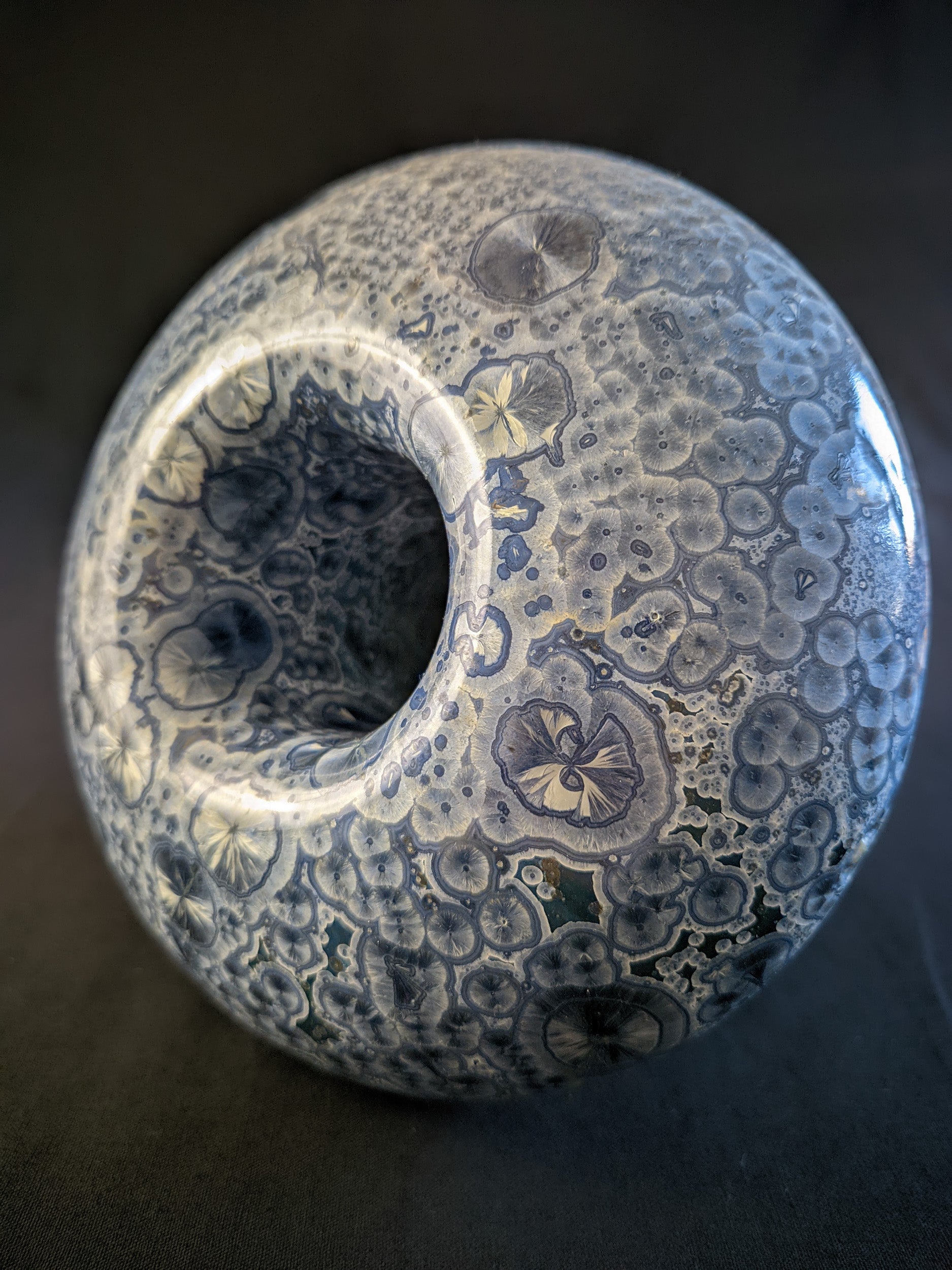 Top of small porcelain vase titled "Cosmic Fungi," featuring light blue and silver ringed crystals, deep blue stem, and a bulbous cap with glaze drips, set against a contrasting background.