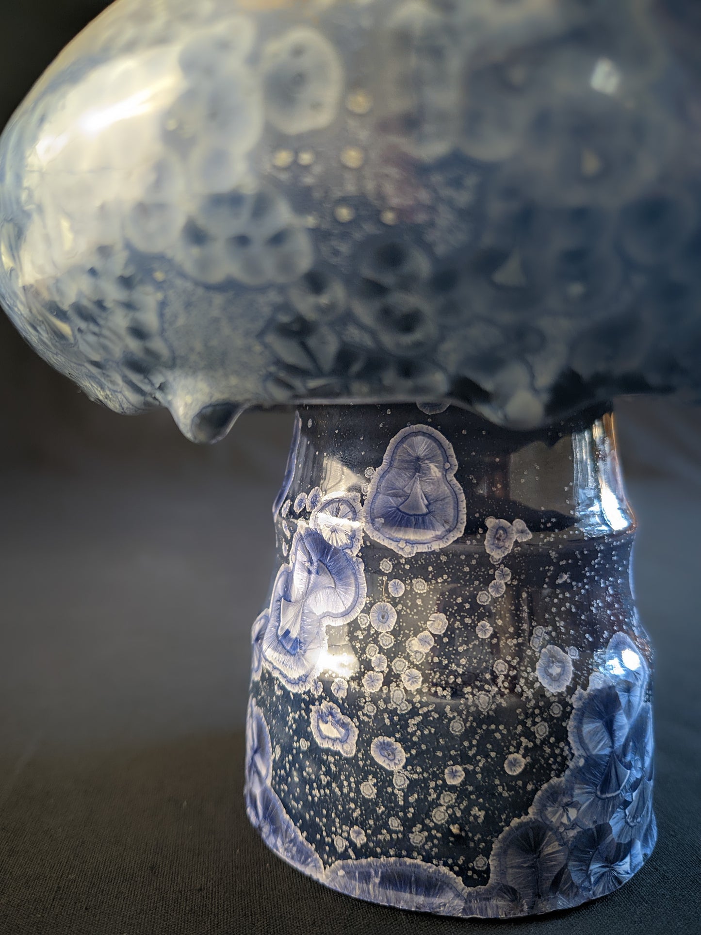 Small porcelain vase titled "Cosmic Fungi," featuring light blue and silver ringed crystals, deep blue stem, and a bulbous cap with glaze drips, set against a contrasting background.