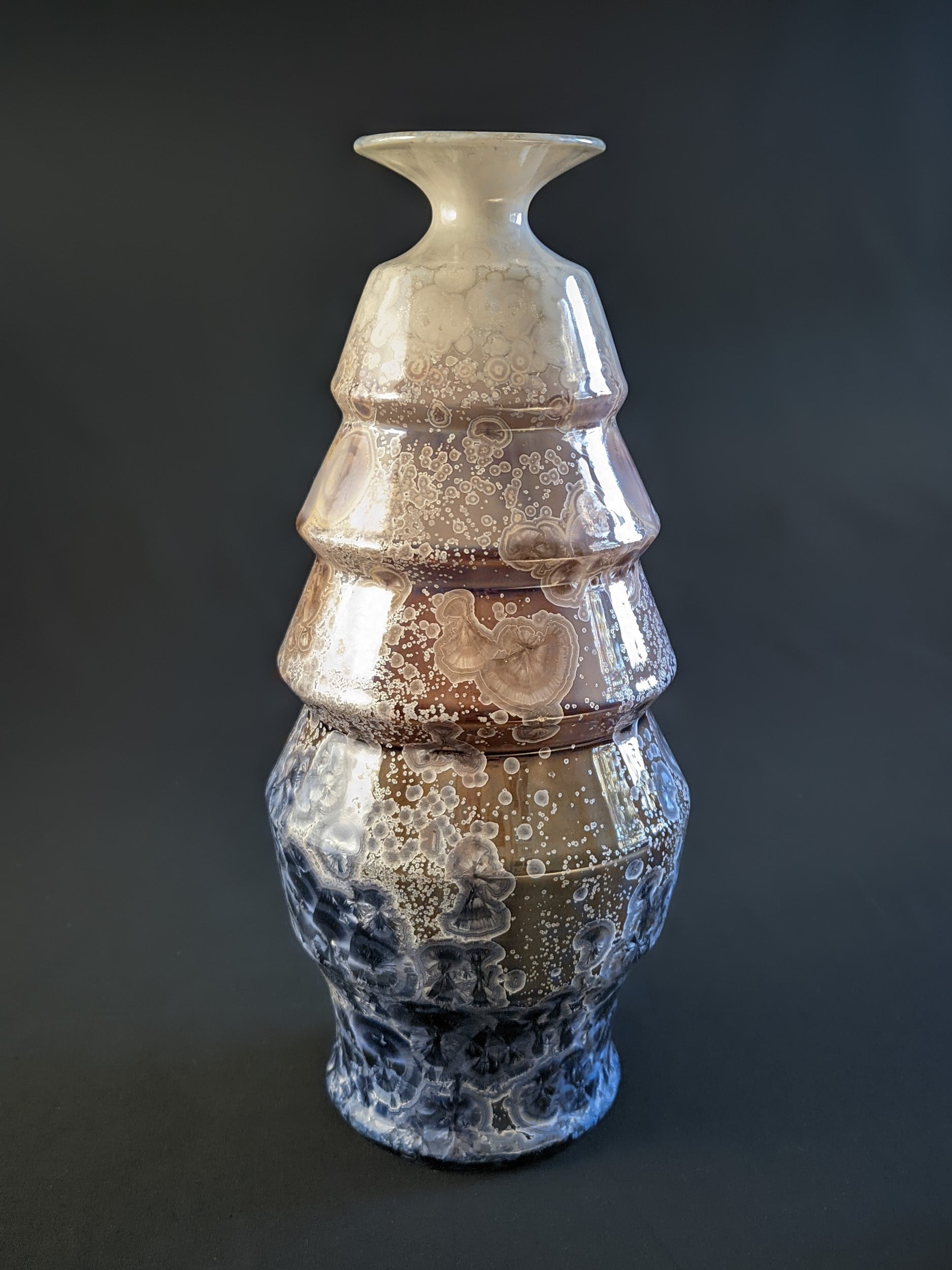 A stunning crystalline-glazed vase named Celestial Cascade, with a tiered, faceted form and intricate starburst-like crystalline patterns in shades of ivory, bronze, blue, and gray.