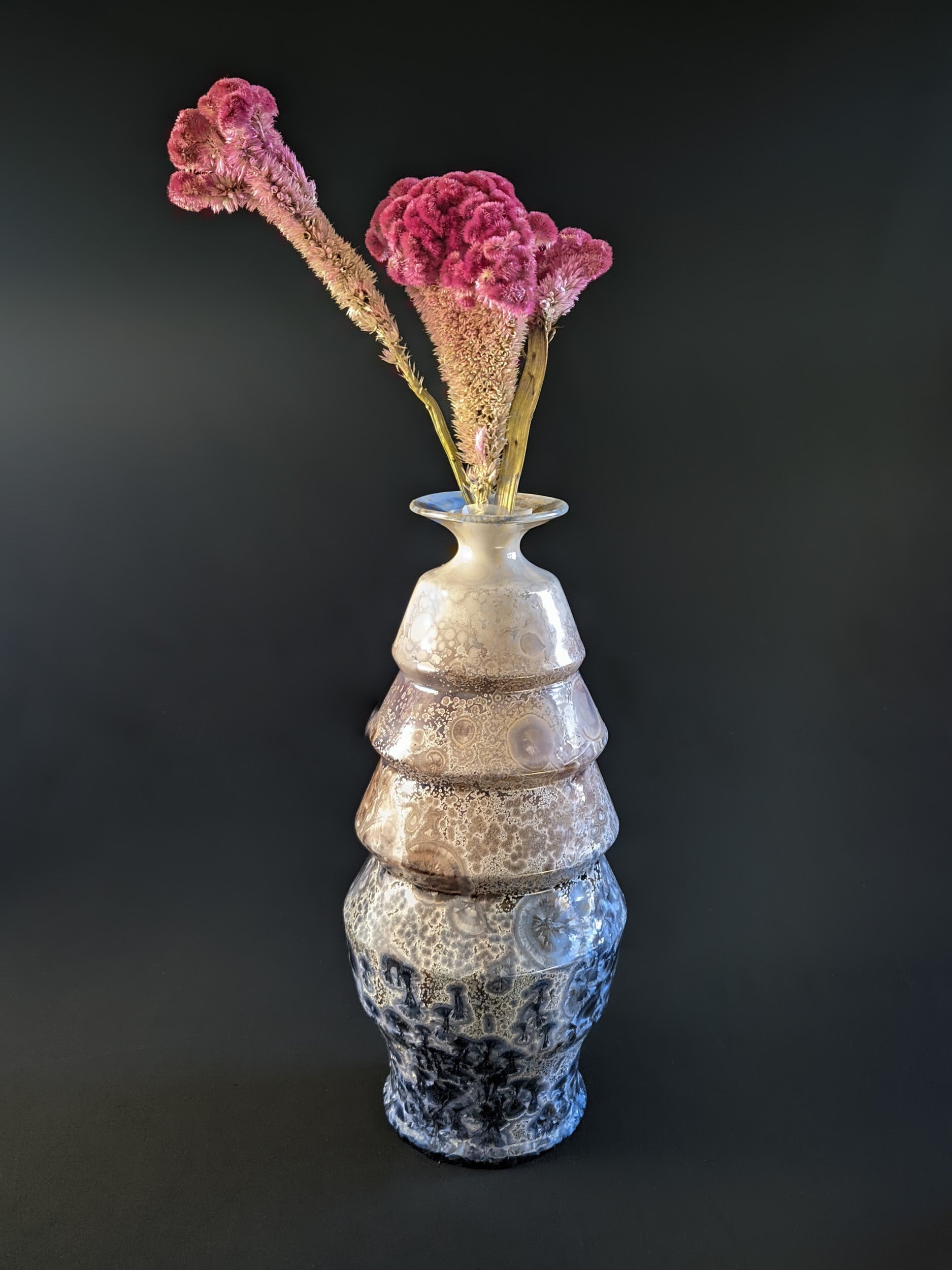 A stunning crystalline-glazed vase named Celestial Cascade, with a tiered, faceted form and intricate starburst-like crystalline patterns in shades of ivory, bronze, blue, and gray showcased with an unusual bright pink flower.