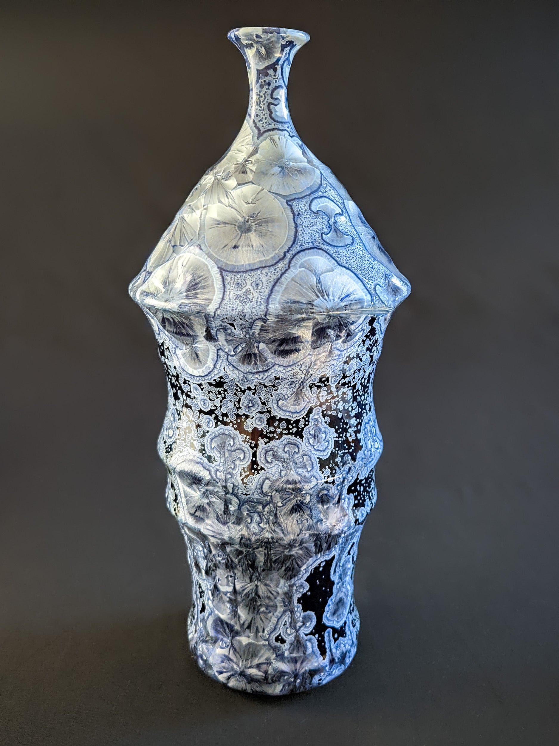 Bottle-shaped ceramic vase with a narrow neck and curved body. Dark blue background with large icy blue crystals and smaller secondary crystals, creating a striking visual effect.