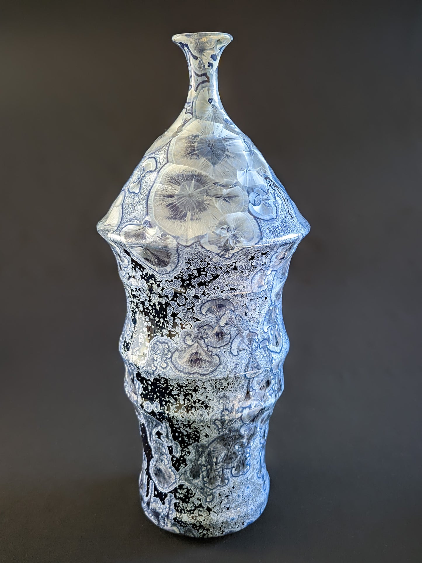 Bottle-shaped ceramic vase with a narrow neck and curved body. Dark blue background with large icy blue crystals and smaller secondary crystals, creating a striking visual effect.