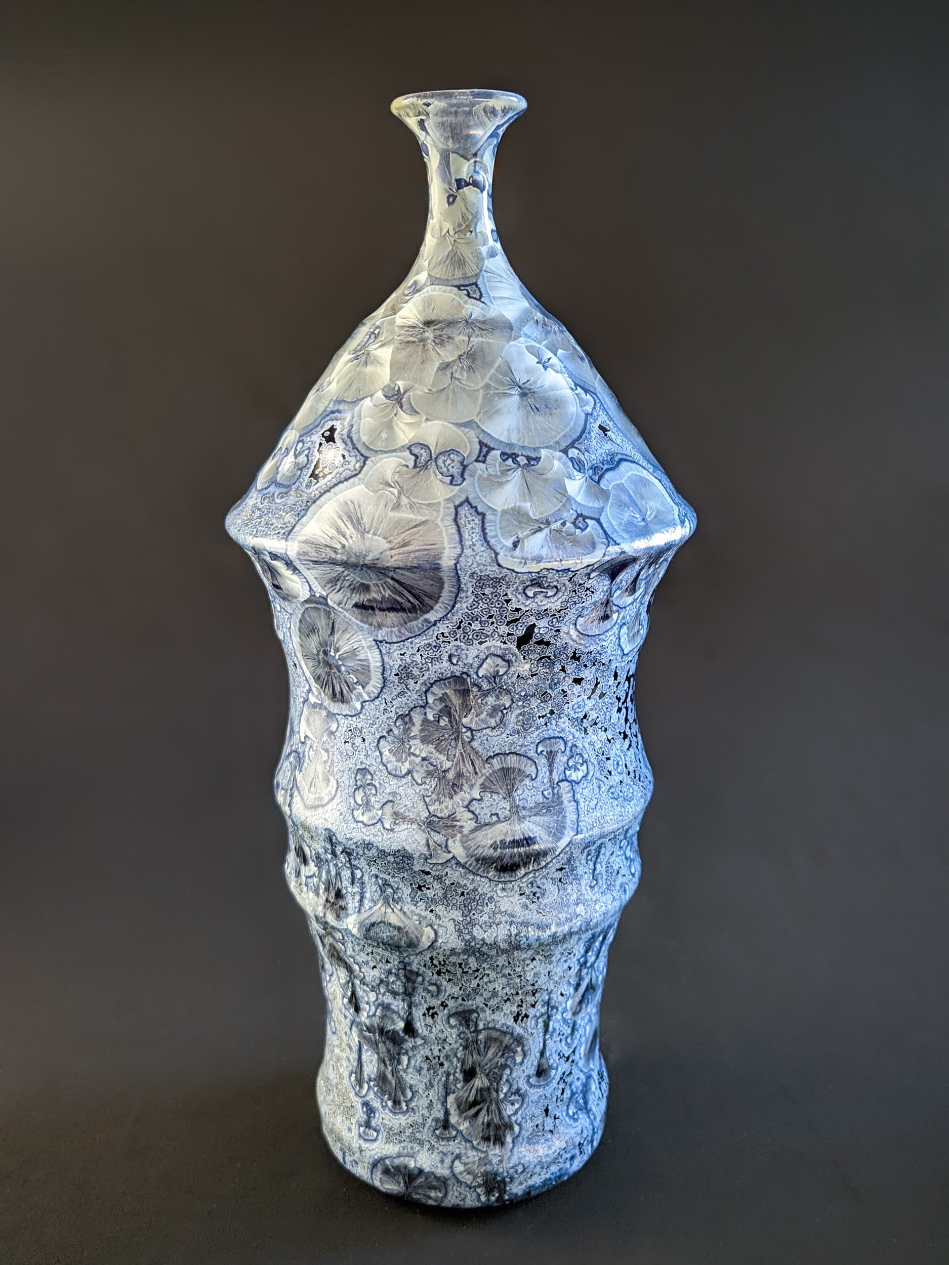 Bottle-shaped ceramic vase with a narrow neck and curved body. Dark blue background with large icy blue crystals and smaller secondary crystals, creating a striking visual effect.