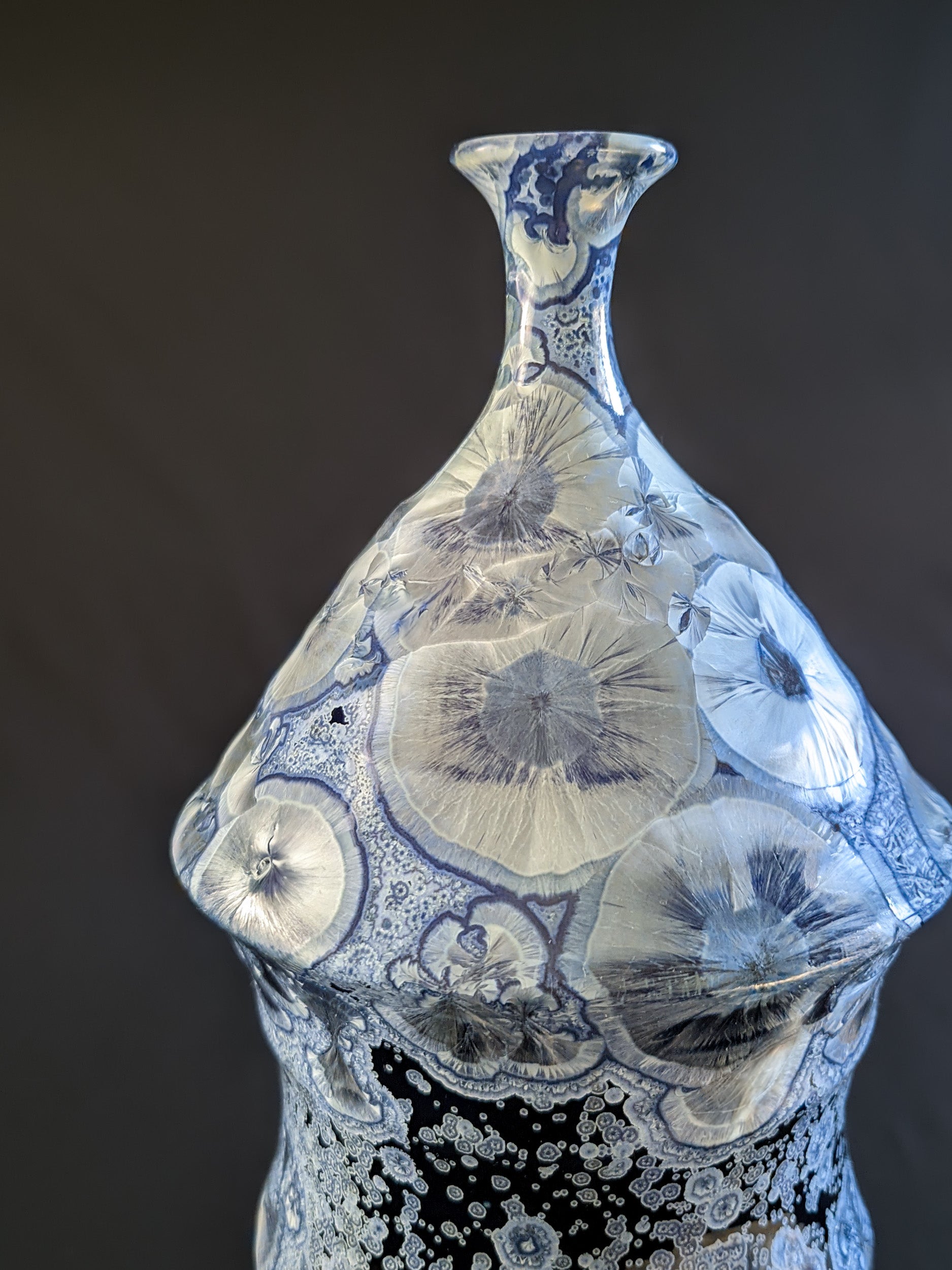Close up of the top of bottle-shaped ceramic vase with a narrow neck and curved body. Dark blue background with large icy blue crystals and smaller secondary crystals, creating a striking visual effect.