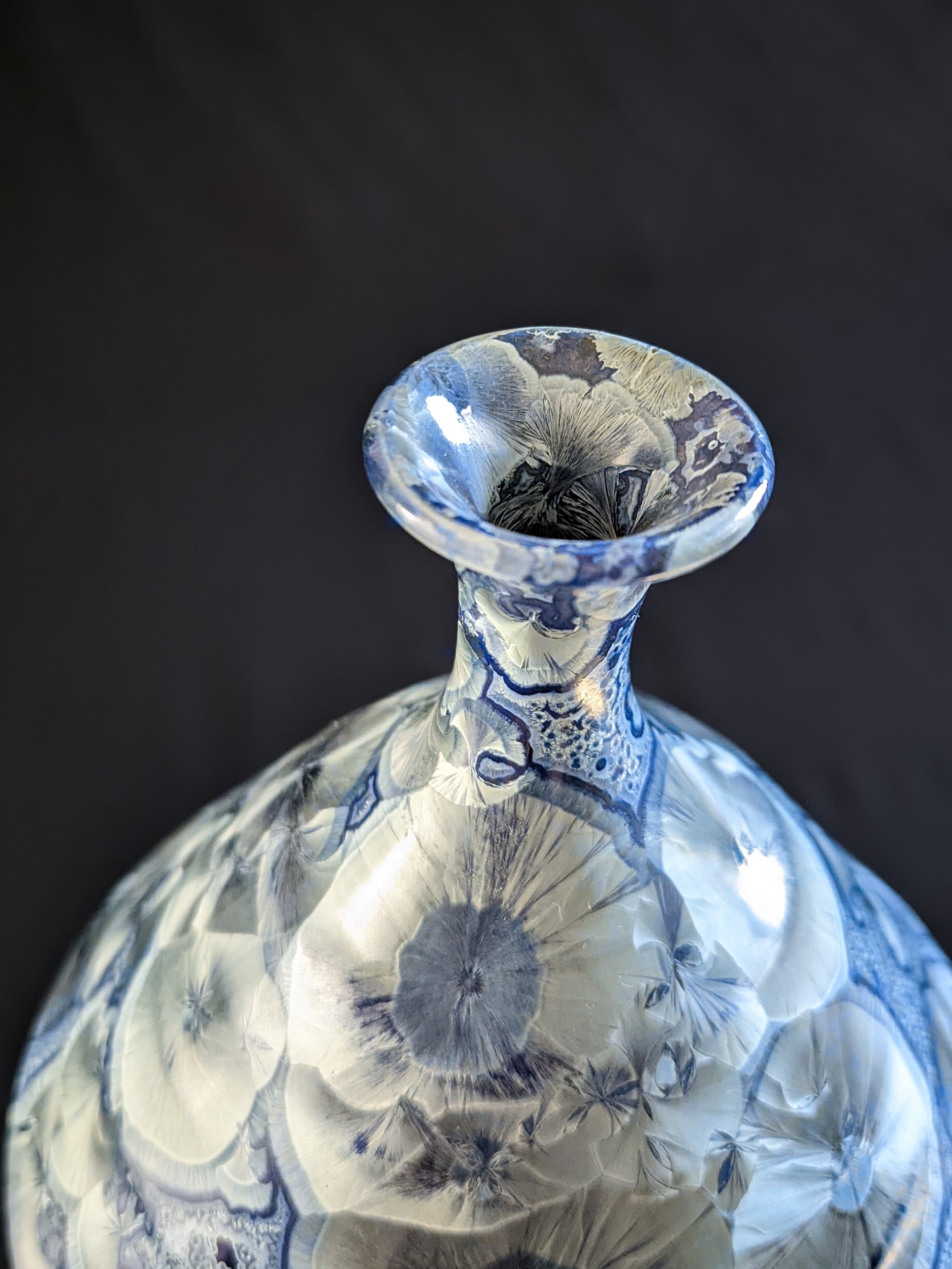 Close up of the top of bottle-shaped ceramic vase with a narrow neck and curved body. Dark blue background with large icy blue crystals and smaller secondary crystals, creating a striking visual effect.