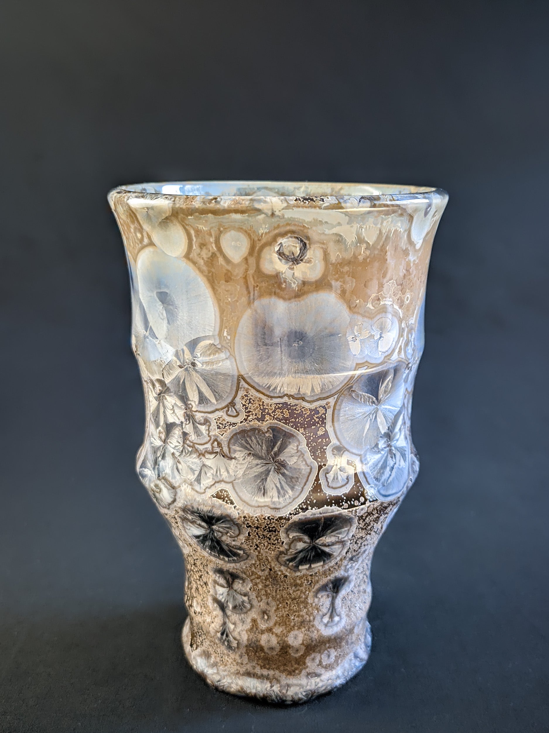 A sculptural vase with a warm amber-brown background, adorned with metallic blue crystalline formations, showcasing a seductive and curvaceous silhouette.