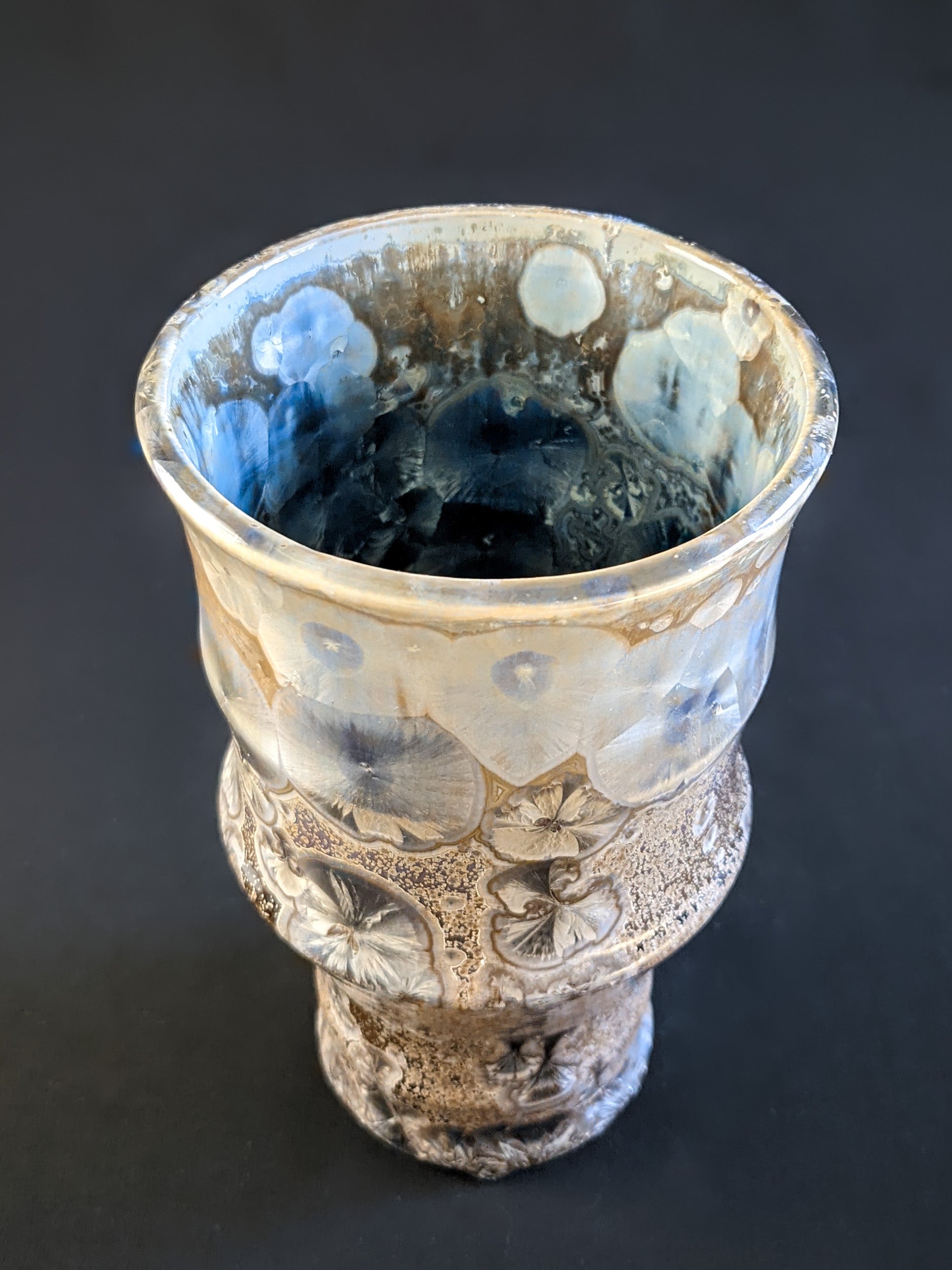 A sculptural vase with a warm amber-brown background, adorned with metallic blue crystalline formations, and a peek at the saphire crystals on the inside. 