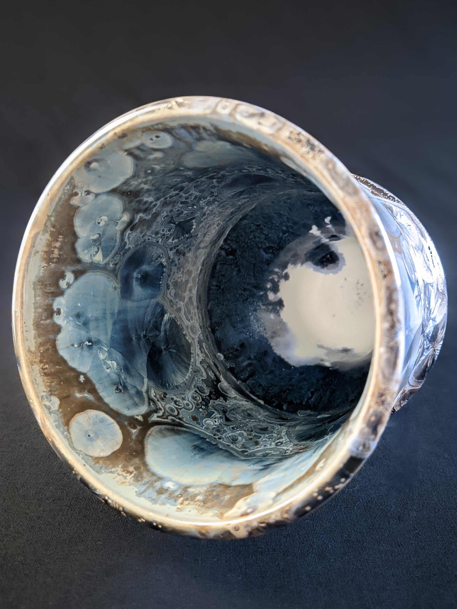 Looking down at the top of the sculptural vase, revealing its wide opening and unique curvature, with a burst of metallic blue crystals forming an intricate pattern against the amber background.