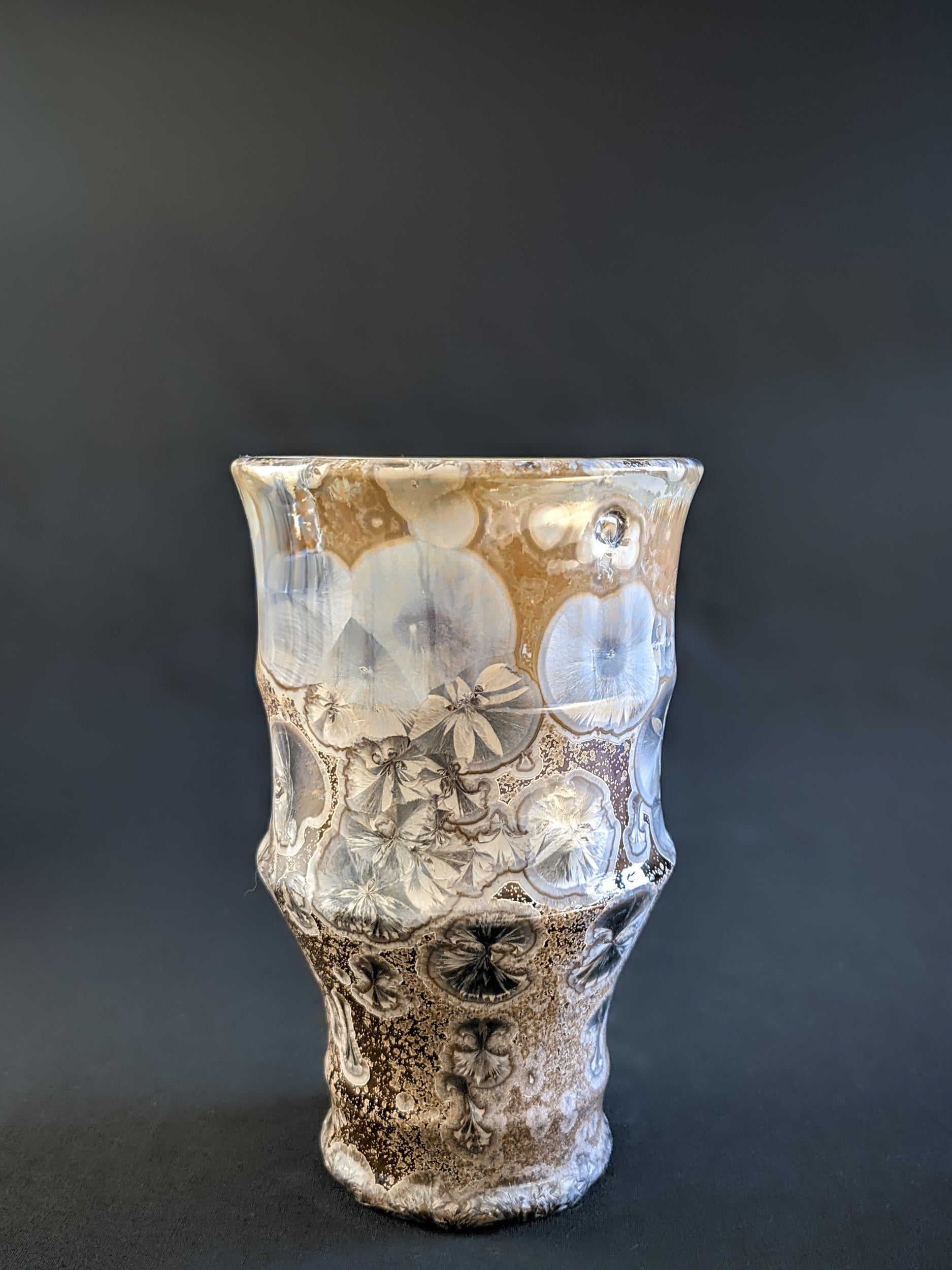 A sculptural vase with a warm amber-brown background, adorned with metallic blue crystalline formations, showcasing a seductive and curvaceous silhouette.