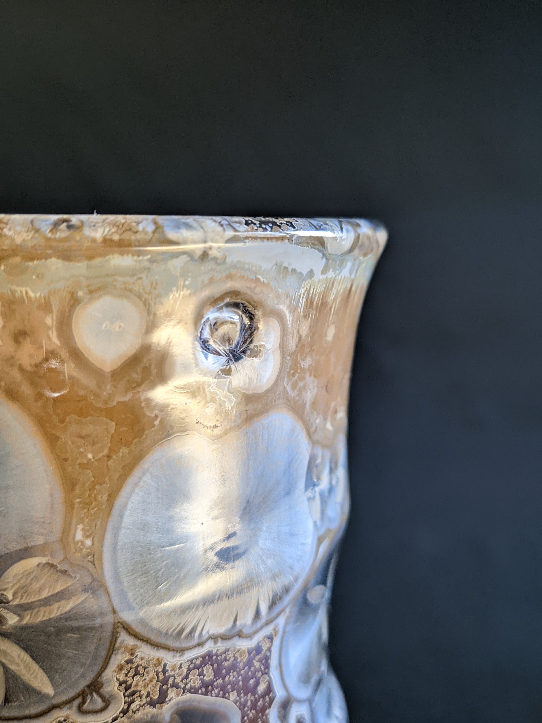 A close-up of the rim of a sculptural vase, revealing its smooth curves and the delicate transition from amber to metallic blue crystals.