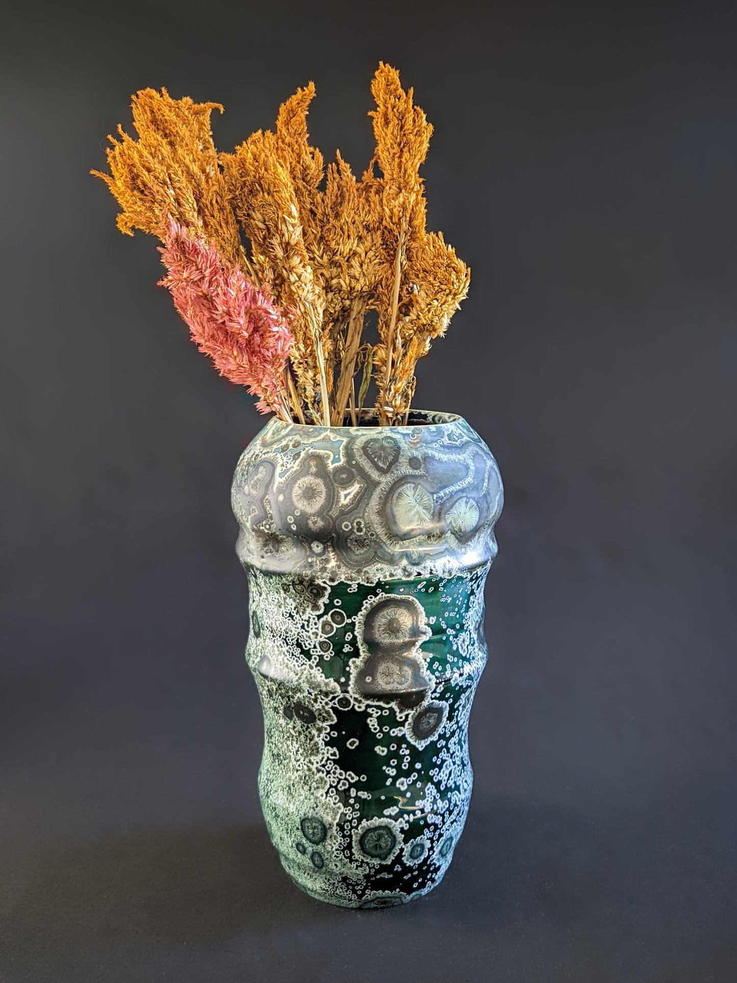 Emerald green crystalline-glazed vase filled with ochre-colored dried flowers, highlighting its sculpted form and intricate gold and nickel crystal patterns.