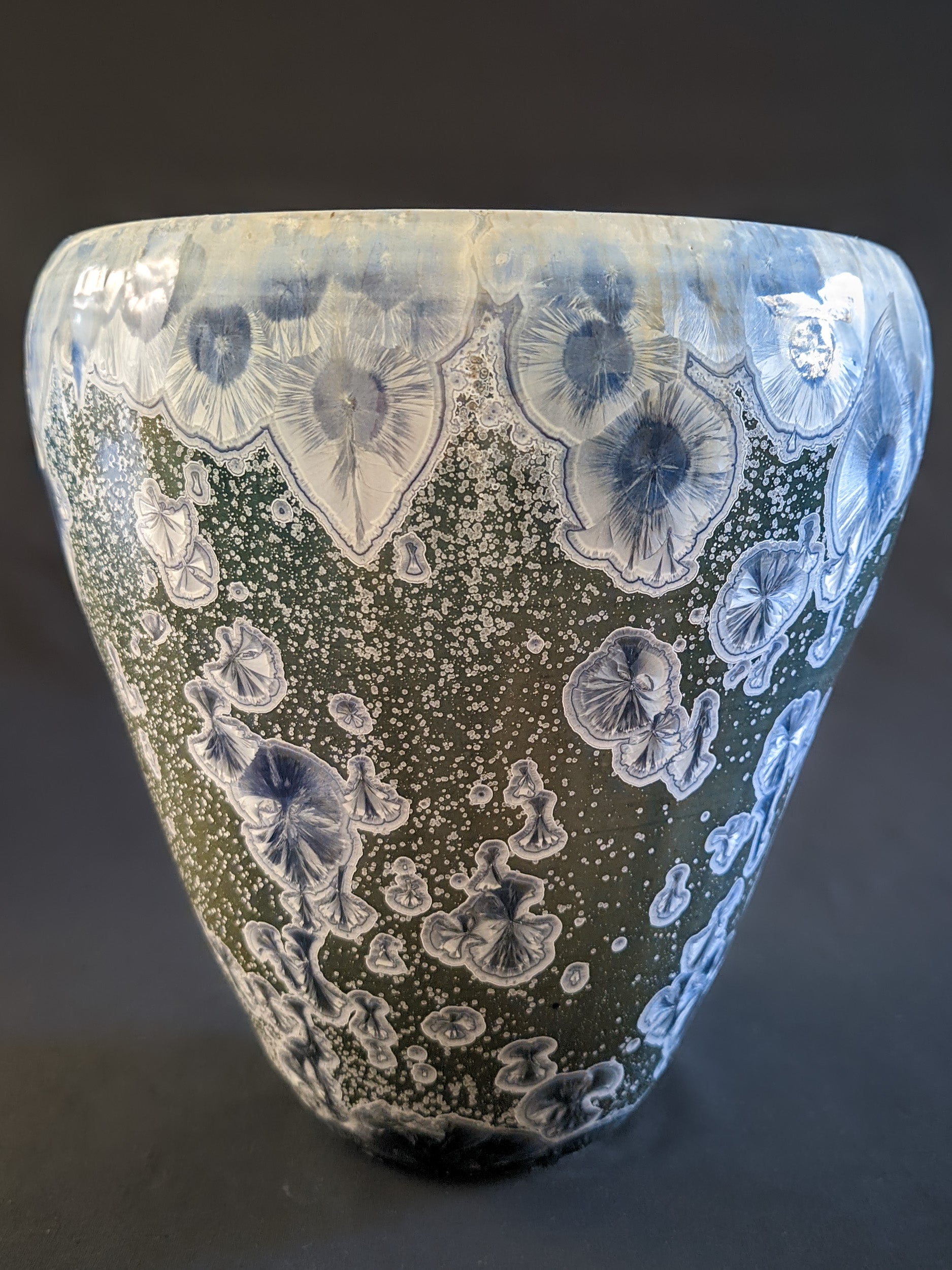 Front view of the sculptural vessel with a muted green background, featuring large deep blue crystals that gradually transition to light blue, accented by a white ring, and thousands of small secondary white crystals covering the surface.